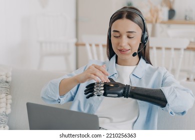 Female Student With Disability Wearing Headset Learning At Laptop At Home. Woman With A Disability Setting, Touching Her Modern Bionic Prosthetic Arm Before Work Or Distant Education On Computer.