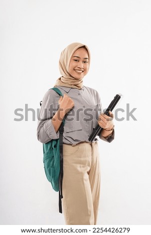 a female student with hijab standing with smile while bring the green bag at her shoulder and the laptop at her hand on the white background