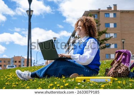 Female student smiling while using laptop and studying outdoors on campus lawn, enjoying pleasant weather and academic pursuits
