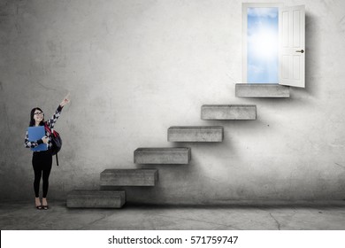 Female student standing on the stairs while pointing at an opportunity door to success with bright sunlight