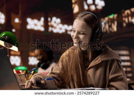 Female student takes notes while she participates at the online remote lesson, using her headset to listen to the tutor. University scholar writes information from the webinar live event.