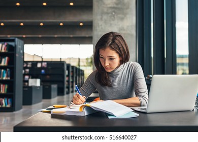 Female student taking notes from a book at library. Young asian woman sitting at table doing assignments in college library. - Shutterstock ID 516640027