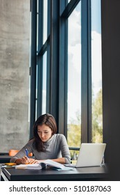 Female Student Taking Notes From Books For Her Study. Young Woman Sitting At Table With Books At University Library.