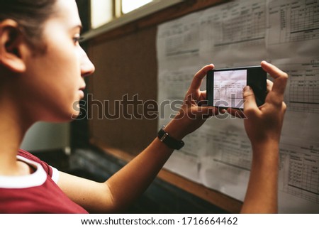 Female student talking a photograph of the notice board in high school. Girl taking a picture of exam timetable with her mobile phone in school.