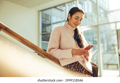 Female Student Using Her Cell Phone While Standing At Steps In University. Young Woman Texting On Mobile Phone In College Campus.