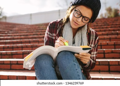 Female Student Using Marker Pen To Highlight Text In A Book. Woman Student Sitting On Steps At Campus And Studying.