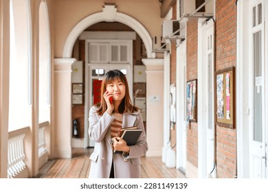 female student wearing stylish grey overcoat chatting over phone while reading a book in university campus in Hong Kong - Powered by Shutterstock