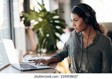 Female Student Working On Laptop With Headphones For A University Project In A Cafe, Restaurant Or Coffee Shop. Thinking And Studying Young University Or College Woman Typing And Listening To Music
