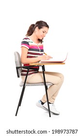 Female Student Writing In A Notebook Seated On A School Desk Isolated On White Background