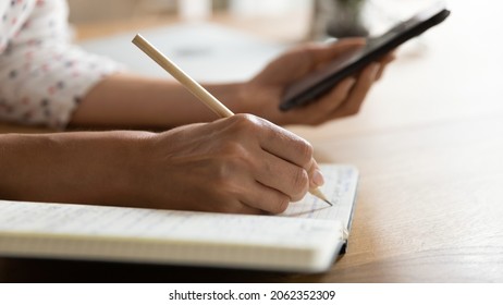 Female student writing in notebook, watching learning webinar on smartphone, preparing for test. Hands of woman taking notes, using online organizer app on cellphone for planning work tasks. Close up