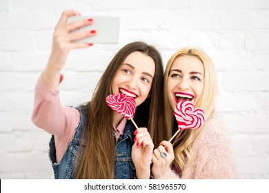 Female stylish friends making selfie photo with red candies on the white wall background