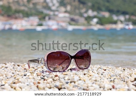 Female sunglasses on pebble stones on sea background. Beach vacation in summer, defocused view to Mediterranean resort