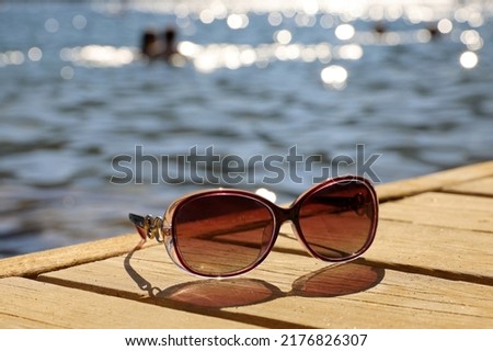 Female sunglasses on wooden boards on sea and swimming people background. Beach vacation in summer, defocused view to shining water