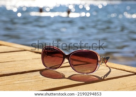 Female sunglasses on wooden boards on sea and swimming people background. Beach vacation in summer, defocused view to shining water