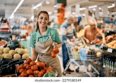 Female supermarket employee arranging fresh tomato at vegetable section and looking at camera. - Powered by Shutterstock