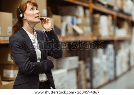 Female supervisor checking boxes on pallets while adjusting headset mic in warehouse, copy space. Logistics, inventory, supply chain, efficiency, industrial, organizational, operational