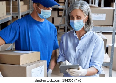 Female supervisor wearing face mask using digital tablet in warehouse talking to male courier holding shipping parcels boxes delivering packages. Covid 19 safety at work and safe shipping delivery.