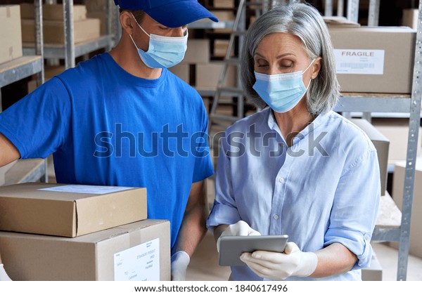 Female supervisor wearing face mask using\
digital tablet in warehouse talking to male courier holding\
shipping parcels boxes delivering packages. Covid 19 safety at work\
and safe shipping\
delivery.