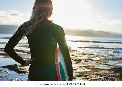 Female Surfer Dressed In Black Wet Suit Is Standing At The Waters Edge With One Hand On Her Hip Looking At The Ocean