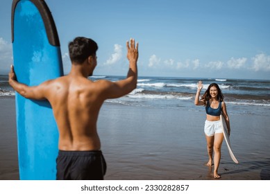 female surfer greets a man with a wave on the beach with her surfboard after practicing - Powered by Shutterstock