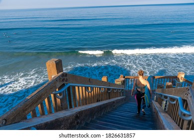 Female Surfer walking down wooden stairs to a san diego beach with a surfboard in her arms while looking out toward the ocean waves.  - Powered by Shutterstock