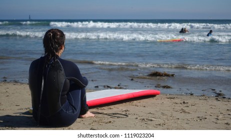 A Female Surfer Wearing A Wet Suit Rests On The Beach And Looks At The Waves On A Beautiful Sunny Day