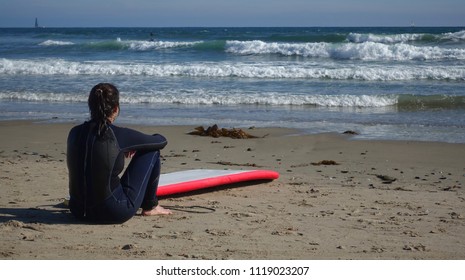 A Female Surfer Wearing A Wet Suit Rests On The Beach And Looks At The Waves On A Beautiful Sunny Day