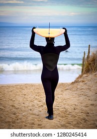 Female Surfer In Wet Suit With Surfboard Balanced On Head Walking Toward The Ocean.