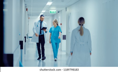 Female Surgeon And Doctor Walk Through Hospital Hallway, They Consult Digital Tablet Computer While Talking About Patient's Health. Modern Bright Hospital With Professional Staff.