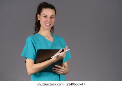 Female Surgeon Smiling In Aquamarine Pre-surgery Scrubs Looking At Camera With Digital Tablet Report On Gray Background