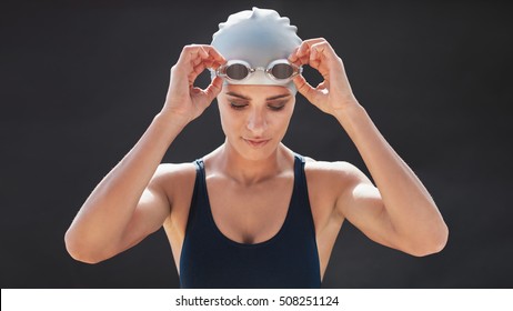 Female swimmer in a swimsuit adjusting her goggles and looking down isolated on black background. Fit young woman in swimming costume. - Powered by Shutterstock