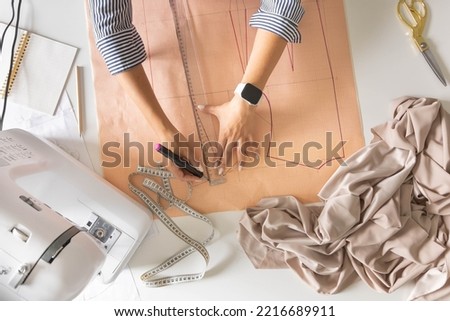 Female tailor hands draw sew pattern ruler and felt tip pen on desk clothes creating workshop top view closeup. Woman dressmaker fashion designer tailoring dressmaking with sewing machine at studio