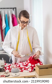 Female Tailor Wearing Glasses Cuts Polkadot Fabric In The Workshop With Scissors.