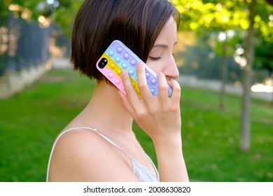Female talking on smartphone with bright pop it bauble case cover in front of green garden. Antistress silicone toy. Simple dimple. Fidget gadget. Autism awareness. Stress reliever