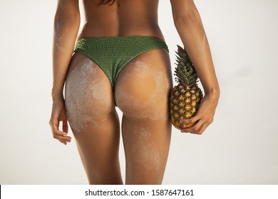 Female Tanned Buttocks In Sand, Sexy Back. Sporty Back Of A Beautiful Woman In Green Bikini With A Pineapple In Her Hands On The Beach Tropical Background With Copyspace.