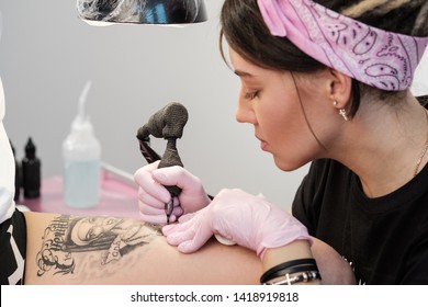 Female Tattoo Master Tattooing Female Client. Figure Nuns On The Thigh. Black Tattoo Machine And Lamp. Workplace Artist. White Background.