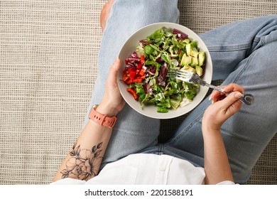 Female with tattoos wearing blue jeans and white shirt sitting and holding a grey bowl with salad and a fork ready to eat, healthy life style concept - Powered by Shutterstock