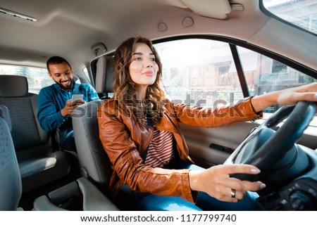 Female taxi driver driving a car with male passenger sitting in the background and using smart phone.