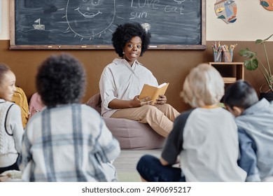 Female teacher of Black ethnicity in bean bag chair reading fairy tale aloud to attentive children sharing thoughts about story sitting around in classroom - Powered by Shutterstock