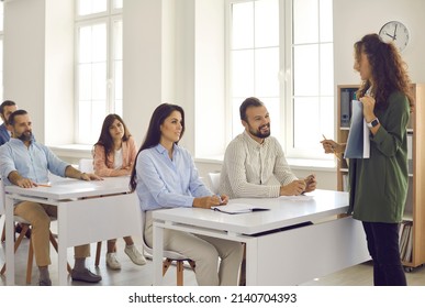 Female Teacher Or Business Coach Communicates With A Group Of Adult Students During A Lecture. People Sit At Desks In The Classroom Taking Advanced Training Courses. Adult Education Concept.