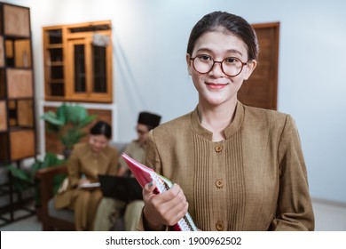female teacher in civil servant uniform wearing glasses smiling while carrying a book with the background of a team of teachers while working from home