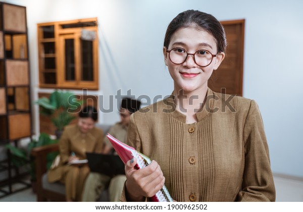 female teacher in civil servant uniform\
wearing glasses smiling while carrying a book with the background\
of a team of teachers while working from\
home