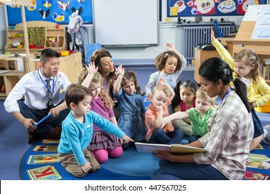 Female teacher giving a lesson to nursery students. They are sitting on the floor and some have their hands up to ask a question.