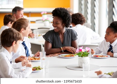 Female Teacher With Group Of High School Students Wearing Uniform Sitting Around Table And Eating Lunch In Cafeteria