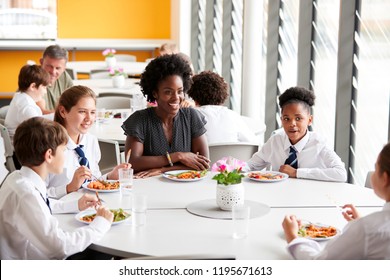 Female Teacher With Group Of High School Students Wearing Uniform Sitting Around Table And Eating Lunch In Cafeteria