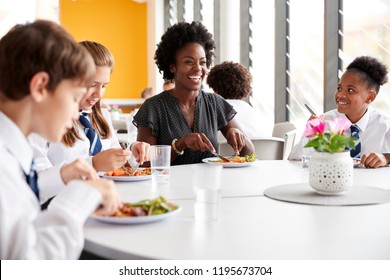 Female Teacher With Group Of High School Students Wearing Uniform Sitting Around Table And Eating Lunch In Cafeteria