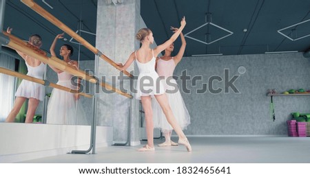 Female teacher shows ballet moves to preteen girl ballerina raising hand by wooden barre in studio at dance lesson