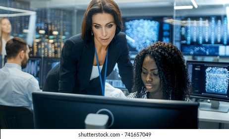 Female Team Leader Consults Young Computer Engineer. They Work In A Crowded Office On A Neural Network/ Artificial Intelligence Project.