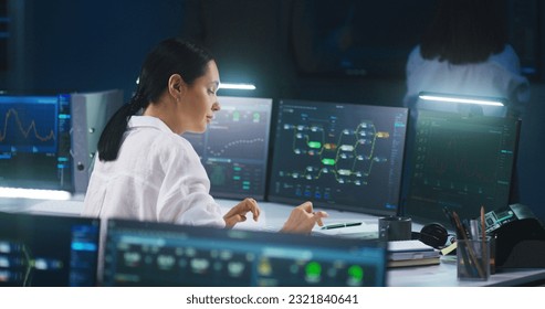 Female IT technical specialist works on computer with data server and blockchain network database in modern monitoring control room. Team of software engineers and big digital screens on background.
