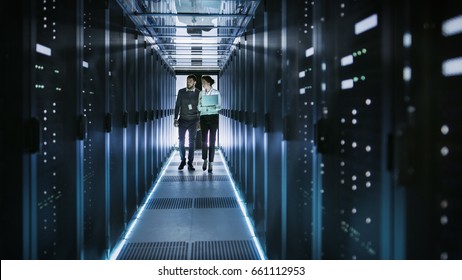 Female IT Technician And Male Server Engineer Talk And Discuss. They Are In Working Data Center Full Of Rack Servers. Woman Holds Laptop.